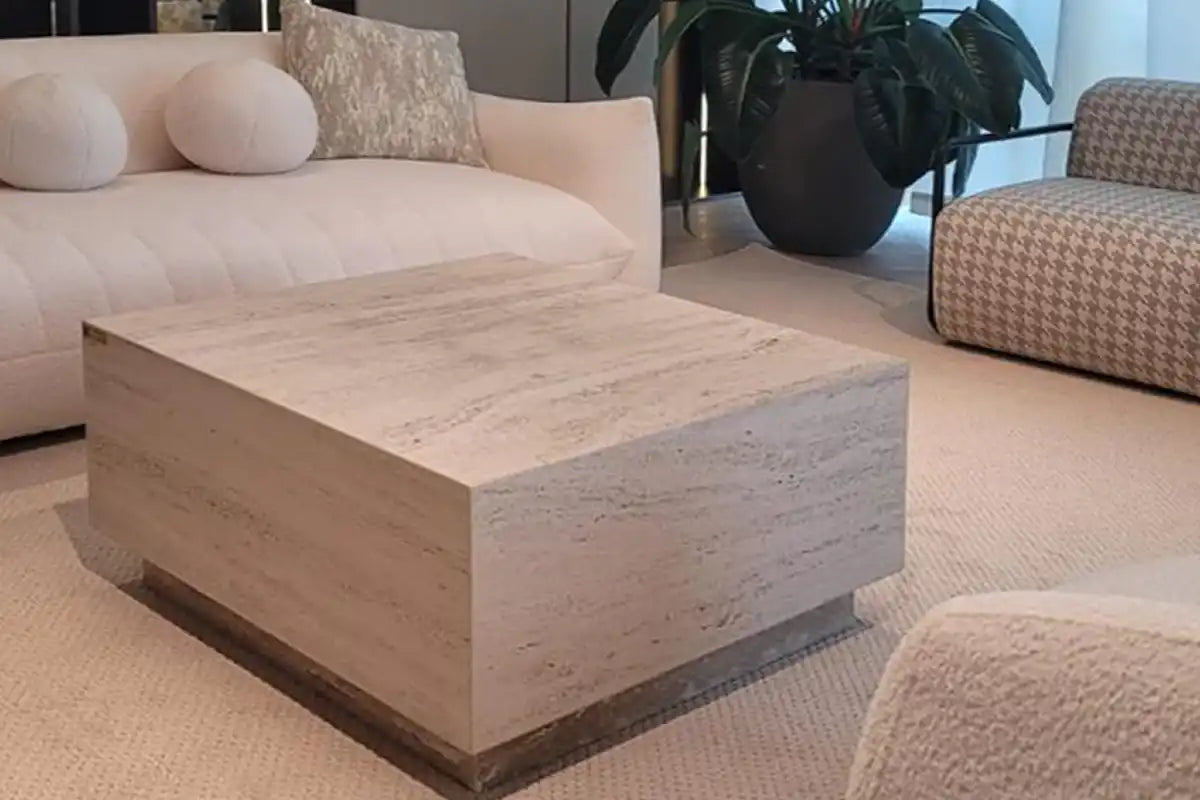 Modern living room with a large square travertine coffee table on beige carpet, surrounded by plush sofas