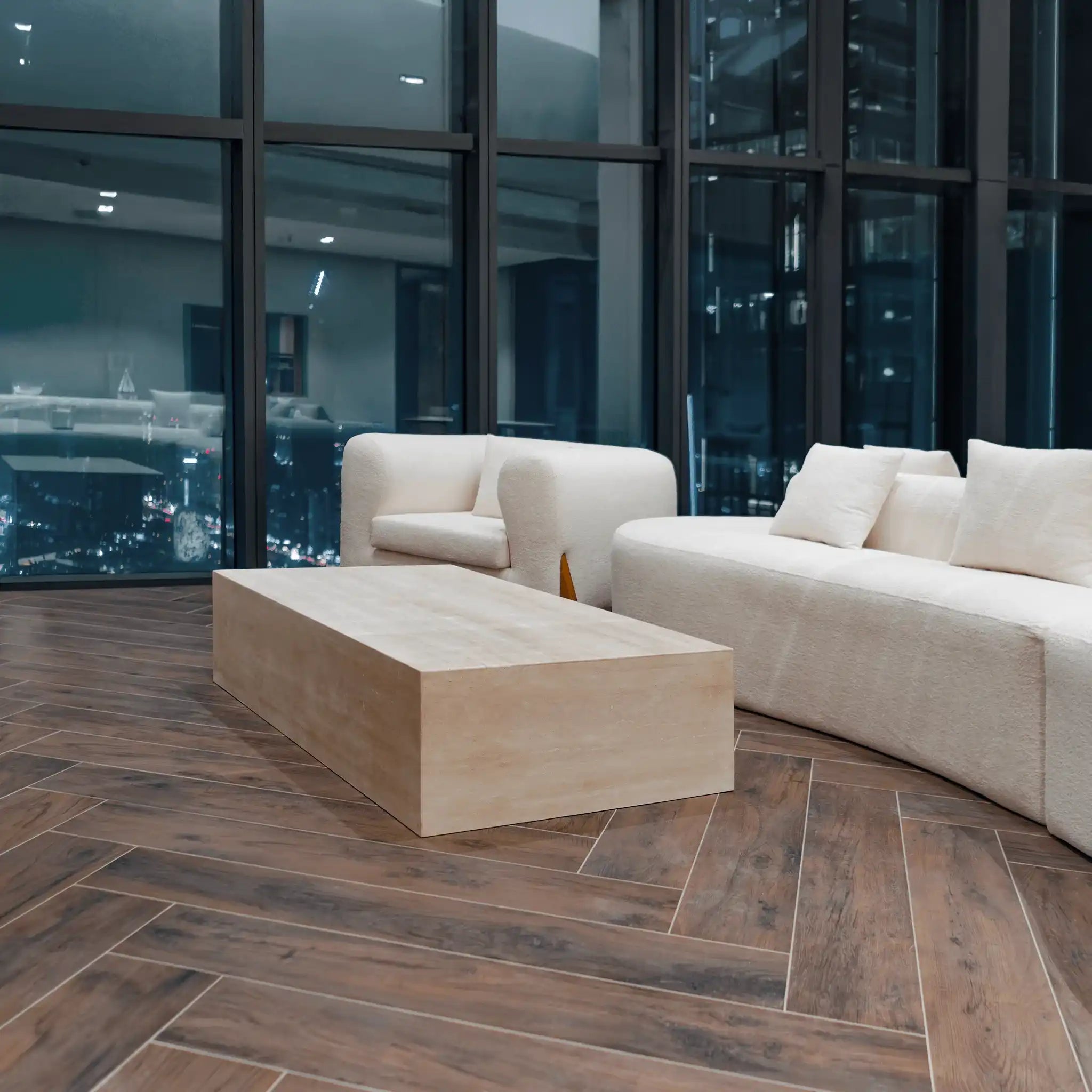 Modern living room with a beige sofa and Linea travertine coffee table from Marbeya Charm in front of large windows with cityscape view.